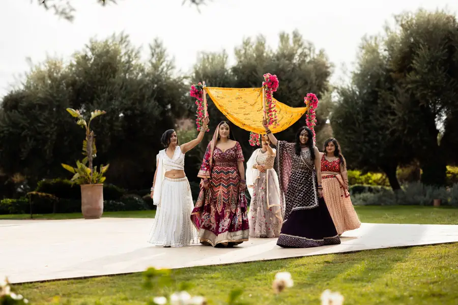 Bridal party in embroidered lehengas carrying a yellow floral canopy across the garden - Indian wedding photographer Marrakech