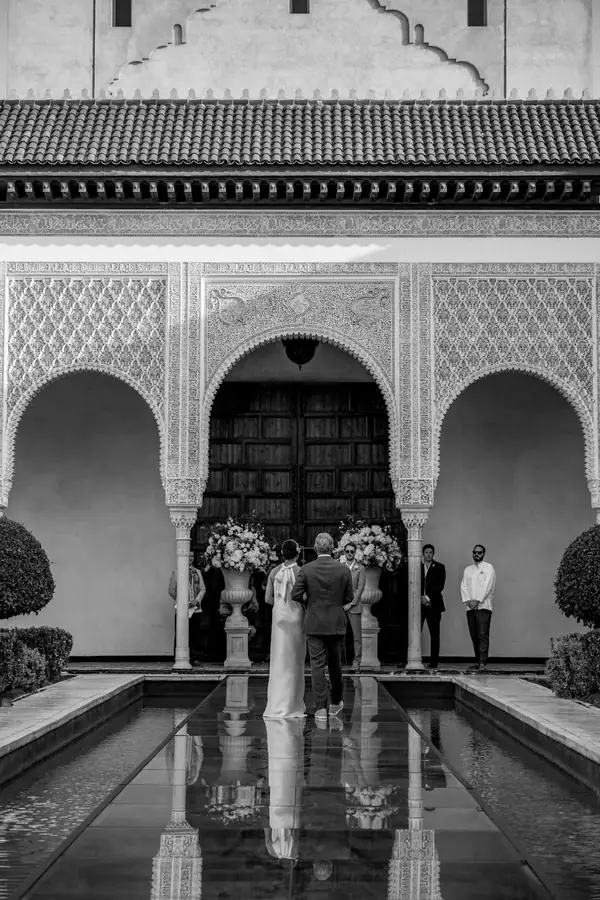 Black and white ceremony before carved Moroccan doors reflected in the courtyard pool - wedding photography Marrakech